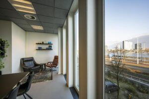 Modern kantoorinterieur aan de De Ruijterkade met zithoek en uitzicht op het IJ en hoge kantoorgebouwen.